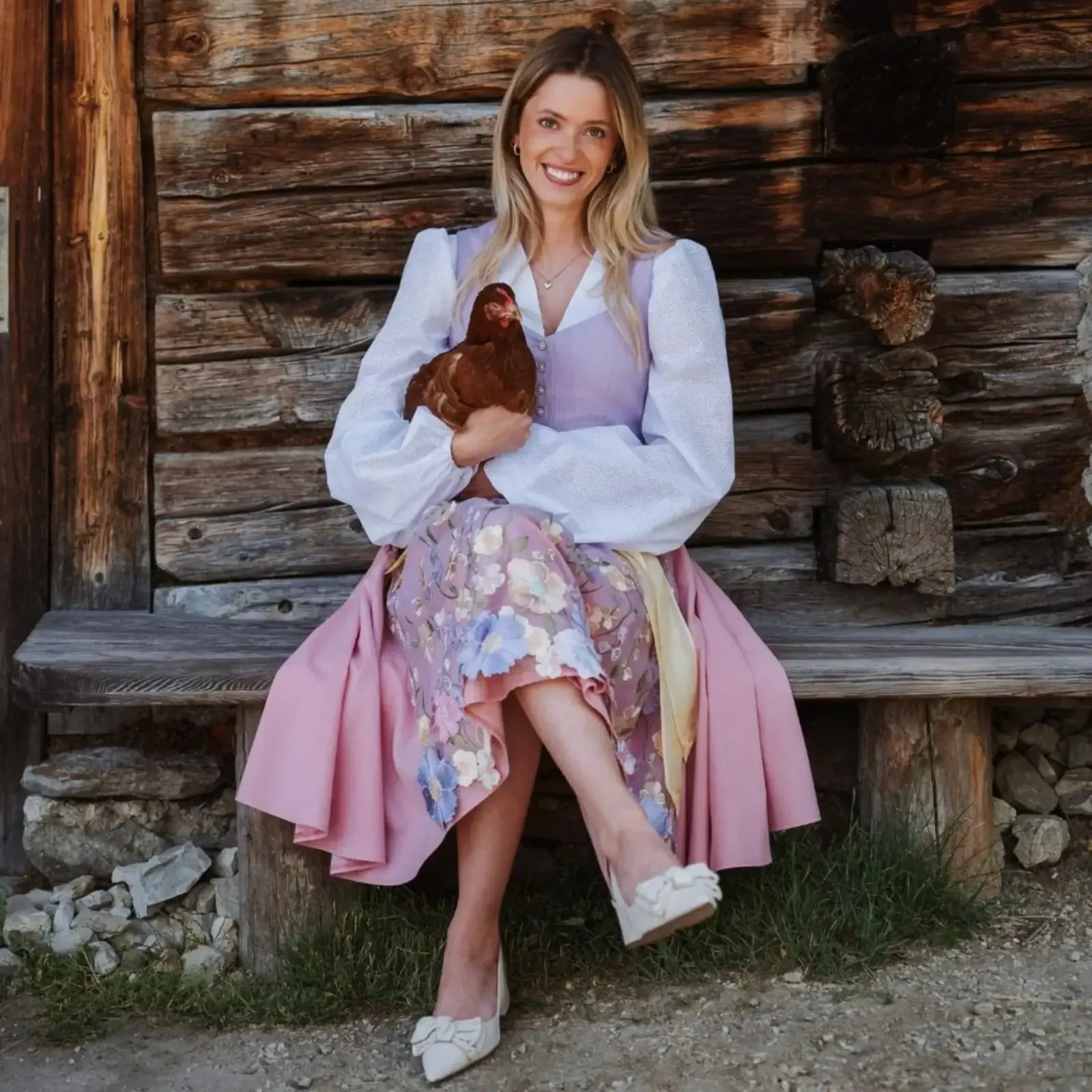 Julia im Dirndl mit einem Huhn im Arm vor einer Holzhütte